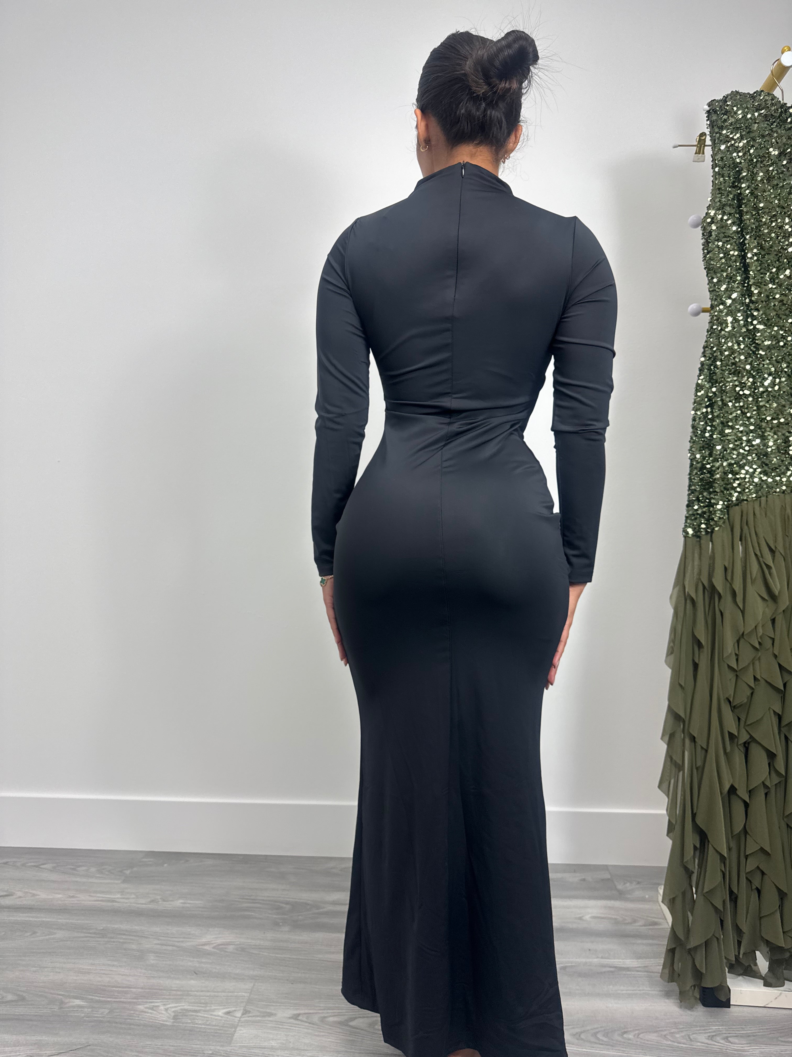 Person wearing a black long-sleeve dress standing in front of a white wall with another dress on a rack to the right.